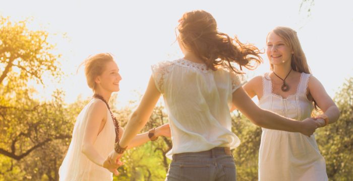 Three young girlfriends dancing in white dresses skipped along the sunset on a meadow at the edge of the forest